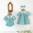Obroi Two turquoise baby dresses with lace collars and matching headbands on wooden hangers against a white wall.