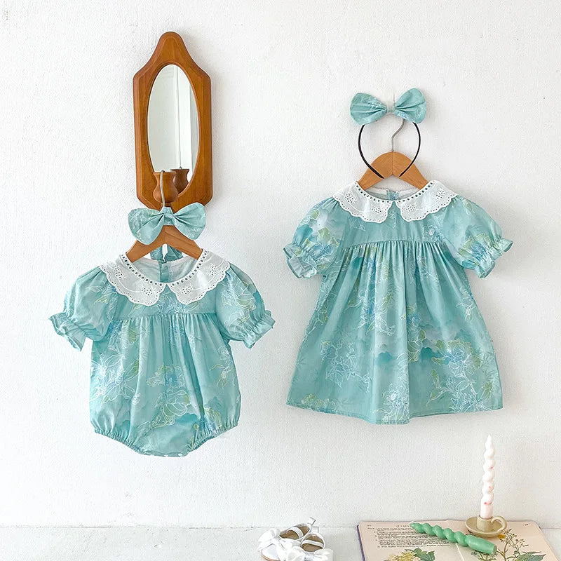 Obroi Two turquoise baby dresses with lace collars and matching headbands on wooden hangers against a white wall.