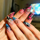 A close-up of hands showcasing long, glossy almond-shaped press-on nails with a variety of colorful designs, including pink, blue, black, and white patterns, decorated with metallic silver glitter stars and rhinestones, Obroi