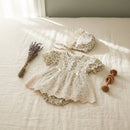 A baby girl's short-sleeve lace romper and matching bonnet laid out on a cream linen bed, styled with dried lavender and a wooden toy, Obroi