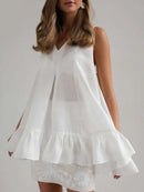 A woman wearing a white sleeveless V-neck blouse with a tiered ruffle hem, paired with a white embroidered mini skirt, against a plain light gray studio background, obroi