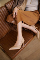 A person seated on a brown leather sofa wearing beige pointed-toe mule heels with a thin stiletto heel and a smooth matte finish, Obroi