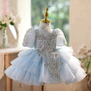 A light blue and white baby girl's party dress displayed on a gold dress form mannequin. The dress features a light blue tulle skirt, a white tulle overlay, and intricate silver beading and sequins on the bodice and sleeves, Obroi