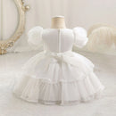 White baby girl's party dress displayed on a dress form mannequin, featuring puff sleeves, a tiered tulle skirt, and a large bow at the back waist, Obroi