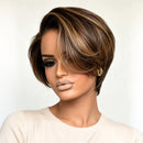 A woman wearing a short, layered wig with dark brown and warm blonde highlights, styled with a side-swept fringe, against a plain light background, Obroi