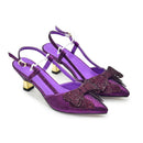 A pair of purple glittery slingback kitten heels with pointed toes, embellished with large matching bow details on the toes and small black fringe, set against a plain white background, Obroi