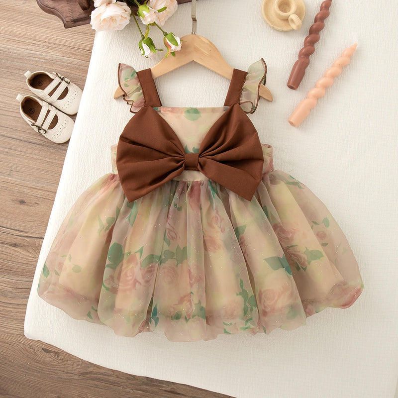 Floral dress with a large brown bow on a white surface, with shoes and toys in the background.