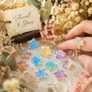 A hand is shown holding a pastel star-shaped resin ring while several matching star charms in pastel colors are scattered on a fabric surface. The scene is styled with dried flowers, greenery, and a small tag reading 'Floral & Stars', Obroi