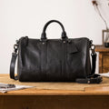 A black leather duffel bag with top handles and a detachable shoulder strap, displayed on a wooden table against a neutral indoor background, Obroi
