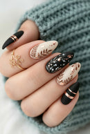 A close-up of a hand showcasing a set of almond-shaped press-on nails. The nails feature a mix of matte black and cream white bases decorated with metallic rose gold foil floral designs, gold stars, and thin metallic bands, Obroi