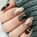 A close-up of a hand showcasing a set of almond-shaped press-on nails. The nails feature a mix of matte black and cream white bases decorated with metallic rose gold foil floral designs, gold stars, and thin metallic bands, Obroi
