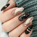 A close-up of a hand showcasing a set of almond-shaped press-on nails. The nails feature a mix of matte black and cream white bases decorated with metallic rose gold foil floral designs, gold stars, and thin metallic bands, Obroi