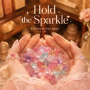 A pair of hands holding a pile of small, sparkling pastel charms in both hands, with a warm, cozy background and the text 'Hold the Sparkle' and 'Charms in your hand.' overlaid at the top, Obroi