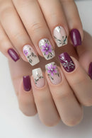 Nails with floral and leaf designs in purple, pink, and white on a neutral background, Obroi
