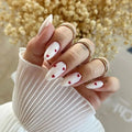 Hand with white nail polish and red heart designs, wearing gold rings, against a blurred background obroi