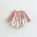 A baby long-sleeve romper with a pink ribbed knit top, a small bow at the neckline, and cream floral bloomers with pink accents, laid flat on a textured white background, Obroi