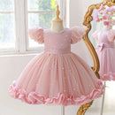 A pink toddler girl's party dress displayed on a mannequin, featuring puff sleeves, a pearl-embellished bodice, and a layered tulle skirt with a ruffled hem. A gold mirror in the background reflects the dress, set against a bright window, Obroi