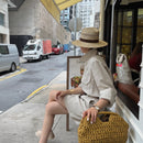 Person sitting on a city street corner holding a woven handbag, wearing a hat and light-colored outfit, Obroi