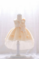 A pale peach and white toddler party dress displayed on a white mannequin dress form. The sleeveless dress features a matching waistband, a large bow on the side, and small floral appliques on the skirt, Obroi