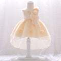 A pale peach and white toddler party dress displayed on a white mannequin dress form. The sleeveless dress features a matching waistband, a large bow on the side, and small floral appliques on the skirt, Obroi