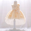 A pale peach and white toddler party dress displayed on a white mannequin dress form. The sleeveless dress features a matching waistband, a large bow on the side, and small floral appliques on the skirt, Obroi