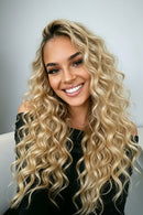 Woman with long, wavy blonde hair smiling in front of a clothing rack, Obroi
