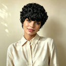 A woman wearing a short, voluminous black curly wig with rose-shaped curls on top, styled with a cream-colored collared blouse against a warm beige background, Obroi