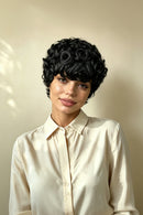 A woman wearing a short, voluminous black curly wig with rose-shaped curls on top, styled with a cream-colored collared blouse against a warm beige background, Obroi