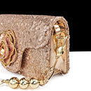 A close-up of a rose gold sequined clutch bag with a gold-tone chain strap, featuring a large fabric rose embellishment on the front flap and polished gold hardware, Obroi