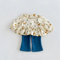 A toddler girl's two-piece outfit featuring a cream-colored long-sleeve ruffled blouse with a green and orange woodland print, paired with blue denim flare pants, laid flat on a light gray background, Obroi