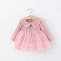 A pink long-sleeve baby dress with a ribbed knit bodice, white scalloped Peter Pan collar, and a layered tulle skirt, hanging on a wooden hanger against a plain white background, Obroi