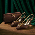 A pair of brown pointed-toe slingback heels with jeweled embellishments, displayed on a wooden surface alongside a matching brown clutch bag against a dark green velvet backdrop, Obroi