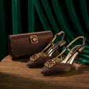 A pair of brown pointed-toe slingback heels with jeweled embellishments, displayed on a wooden surface alongside a matching brown clutch bag against a dark green velvet backdrop, Obroi