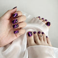 A close-up of a hand and feet showcasing glossy purple nail polish with a metallic cat-eye shimmer effect, styled with draped white satin fabric, Obroi