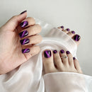A close-up of a hand and feet showcasing glossy purple nail polish with a metallic cat-eye shimmer effect, styled with draped white satin fabric, Obroi