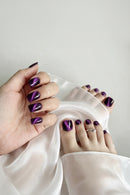 A close-up of a hand and feet showcasing glossy purple nail polish with a metallic cat-eye shimmer effect, styled with draped white satin fabric, Obroi