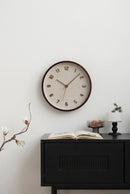 A round wall clock with a textured beige face and dark brown frame, mounted on a white wall above a black cabinet with decorative items, Obroi
