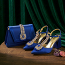 A matching royal blue handbag and a pair of pointed-toe high-heel pumps with ornate gold embellishments and blue jewel accents, arranged on a wooden surface in front of dark green velvet drapery, Obroi