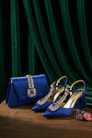 A matching royal blue handbag and pointed-toe high heels displayed on a wooden surface against a dark green velvet curtain. The shoes feature ornate gold embellishments with blue gemstone accents on the toe, straps, and heels, and a gold interior, Obroi