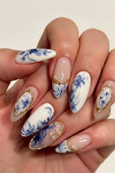 A close-up of hands showcasing an elegant almond-shaped nail art design. The manicure features a mix of glossy white and sheer nude-pink nails decorated with blue floral patterns, delicate gold metallic borders, and small white pearl accents, Obroi