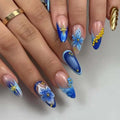 A close-up of a hand showcasing a detailed artificial nail art set. The nails feature various designs including blue and white gradient tips, 3D blue and white flowers, gold glitter starbursts, and gold bead accents on a light gray background, Obroi