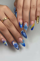 A close-up of a hand showcasing a detailed artificial nail art set. The nails feature various designs including blue and white gradients, 3D blue flowers, gold glitter stars, and gold bead accents on a clean white background, Obroi