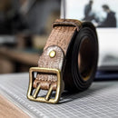 A rolled brown textured leather belt with an antique brass buckle, resting on a gray gridded cutting mat with a blurred workshop background, Obroi