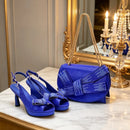 A matching royal blue velvet fashion set featuring a pair of high-heeled slingback sandals and a structured shoulder bag, both adorned with sparkling rhinestone bow details and gold-tone chain straps, displayed on a marble console table in an elegant room, Obroi