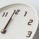 Close-up of a minimalist wall clock with a warm off-white textured face and dark brown wooden numerals and hands, Obroi
