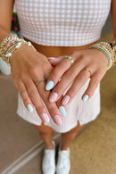 Close-up of a person's hands with pastel nail art designs, including light blue, white polka dots, and white stripes on a pink background. The person is wearing multiple gold rings and pearl/beaded bracelets, Obroi