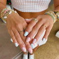 Close-up of a person's hands with pastel nail art designs, including light blue, white polka dots, and white stripes on a pink background. The person is wearing multiple gold rings and pearl/beaded bracelets, Obroi