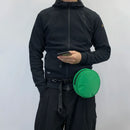 Person wearing a black jacket and holding a green round bag against a white background, Obroi