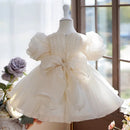 A cream-colored toddler girl's dress displayed on a mannequin from the back, featuring puffy short sleeves, a large bow at the waist, and a voluminous tulle skirt, set against a soft window background, Obroi