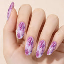 A close-up of a hand with long almond-shaped nails decorated in a purple and pink marble or gemstone-like pattern against a soft beige background, Obroi